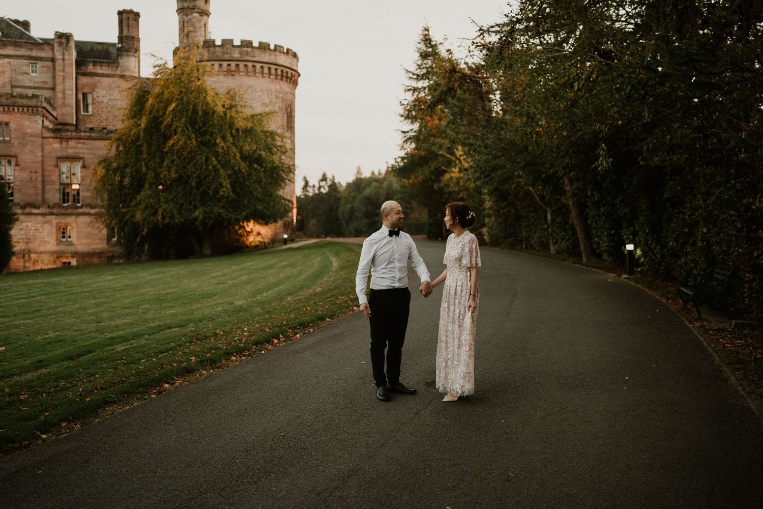 A couple walks along a tree-lined path near a historic castle building at sunset.