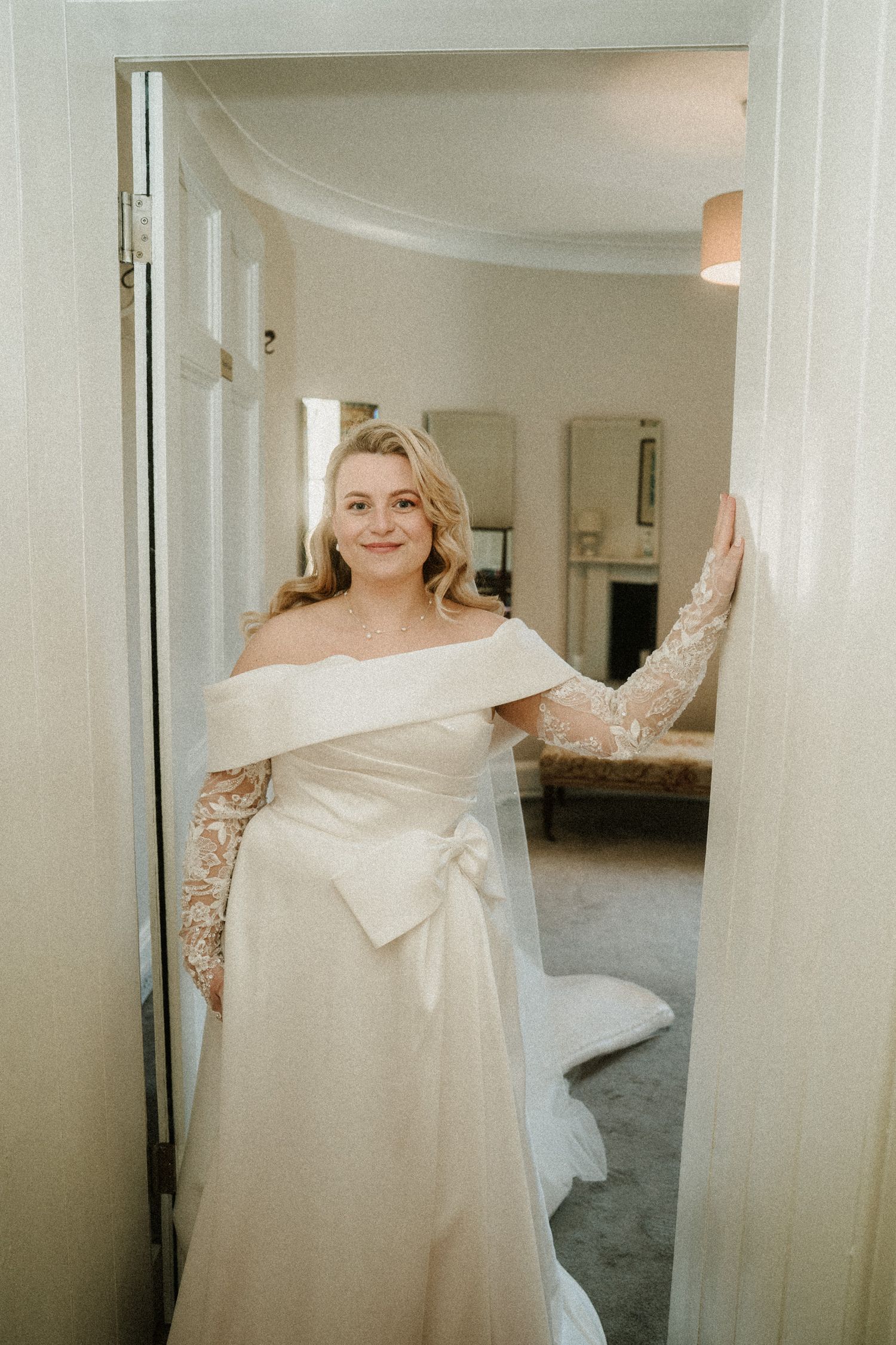A flowing white wedding dress with lace sleeves is displayed in a bright hallway setting.