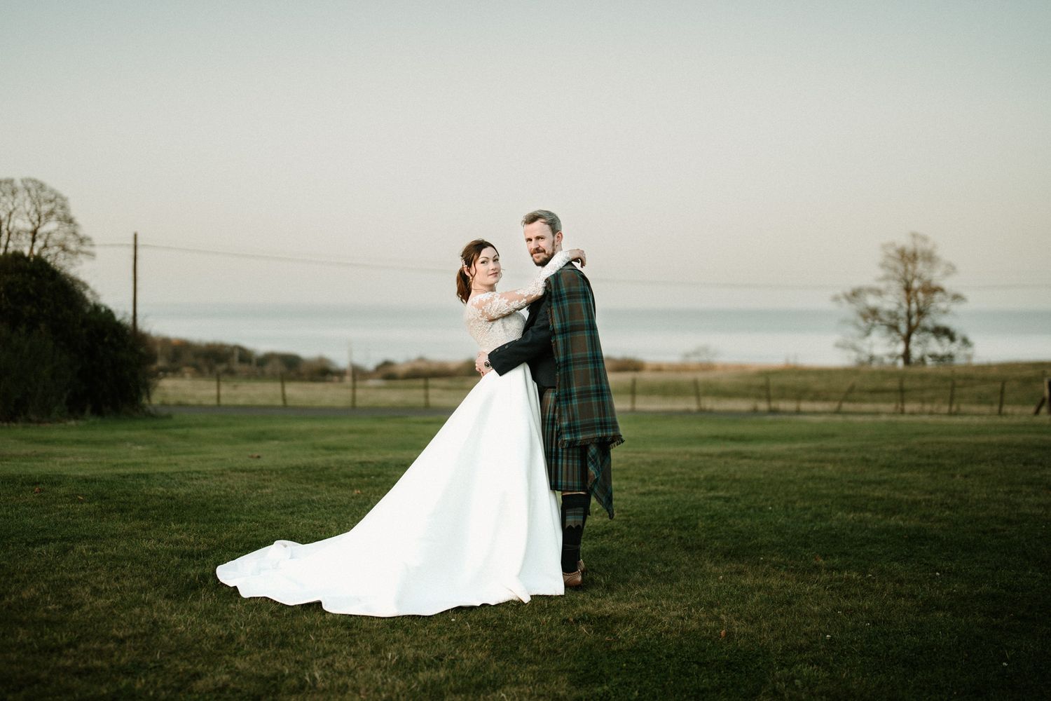 Bride and groom in Scottish attire pose for portraits in an open field.