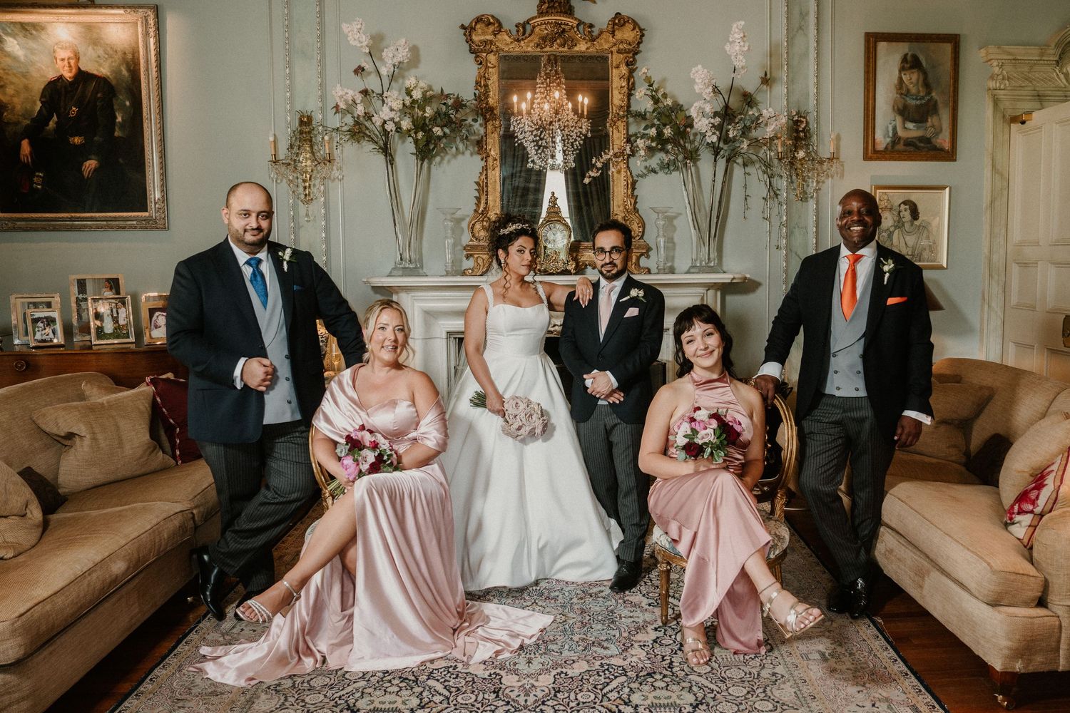 Wedding party in formal attire poses on elegant beige sofa in luxurious room with ornate decor and floral arrangements.