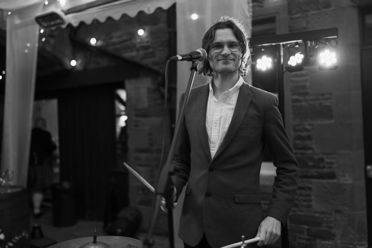 Smiling man in a blazer and open-collar shirt stands at a microphone holding drumsticks on a stage with string lights in the background.