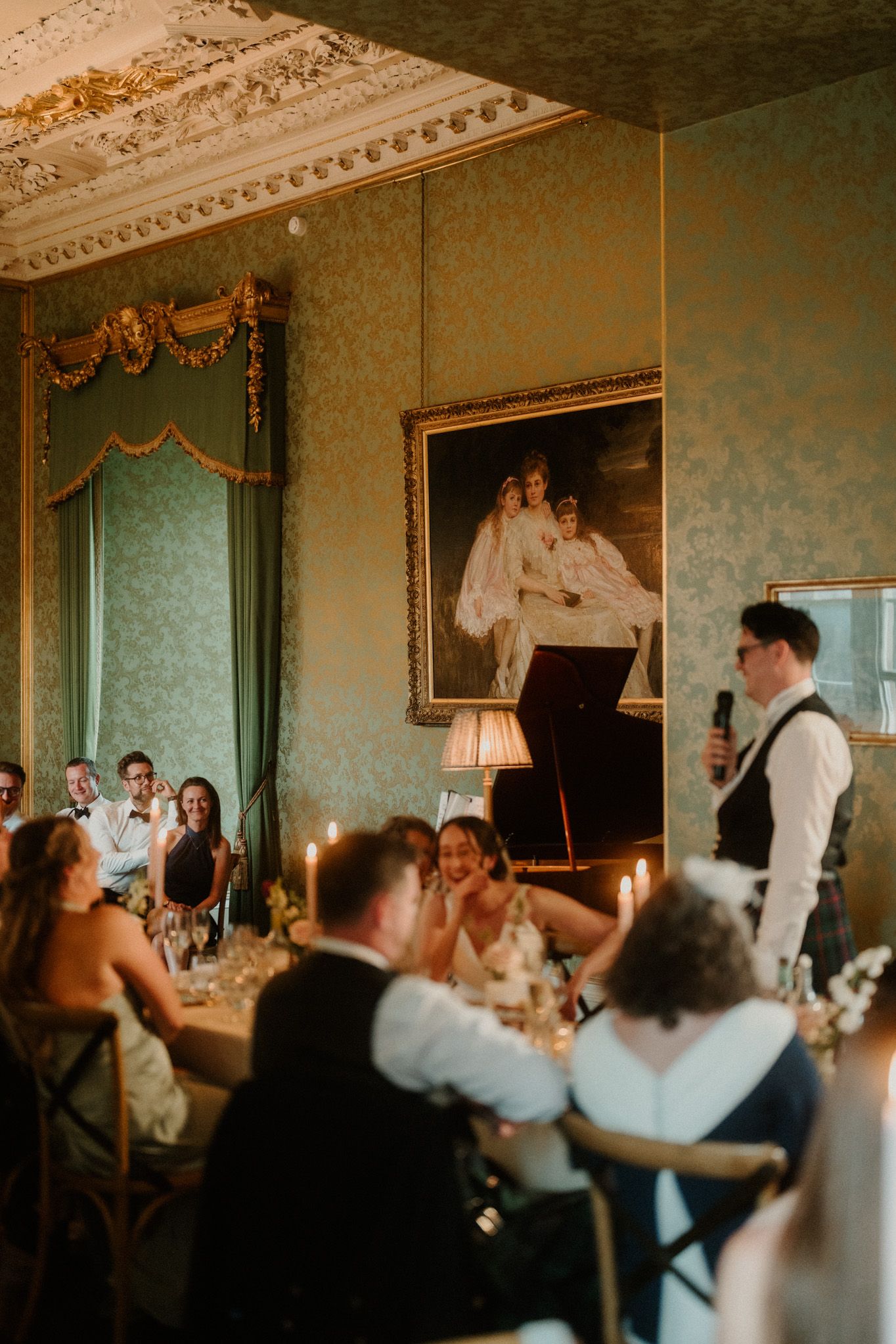 Man in vest gives a wedding speech with a microphone as laughing guests sit at candlelit tables in an ornate green ballroom with a portrait.