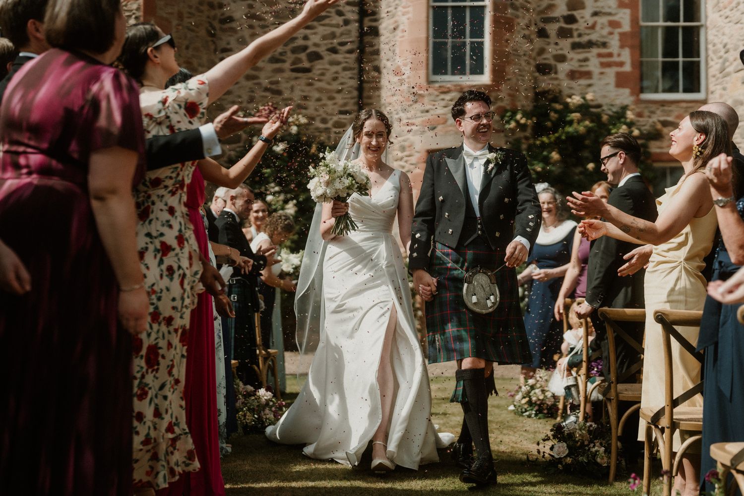 Bride in a white gown and groom in a kilt walk smiling past cheering wedding guests outside a historic stone building.