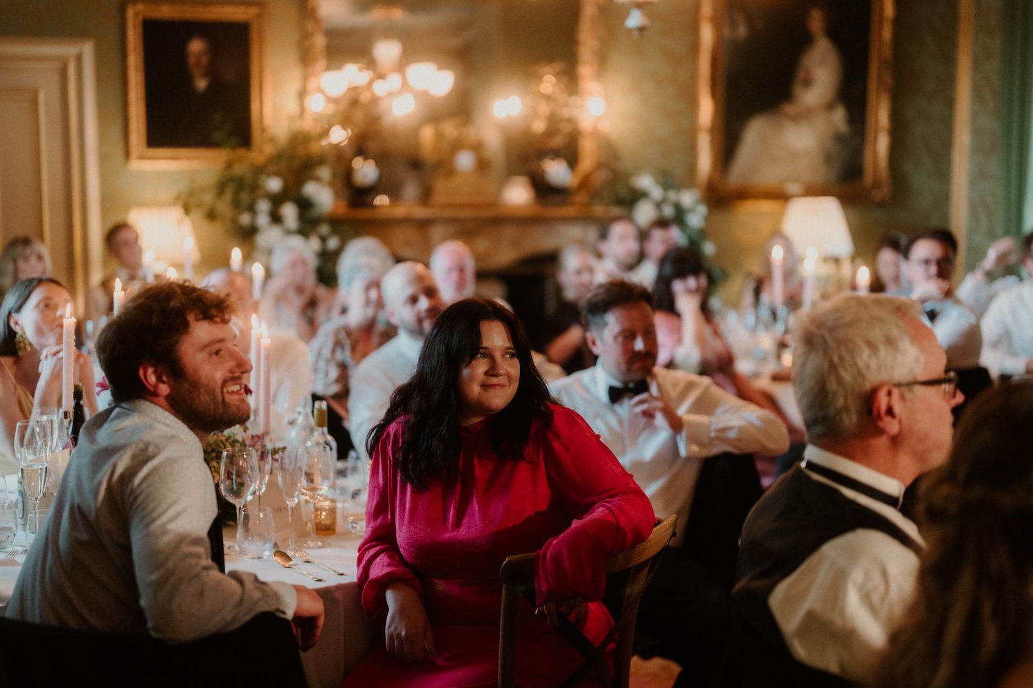 Guests laugh and chat at a candlelit banquet table in an elegant room with framed portraits, with a woman in a pink dress smiling.