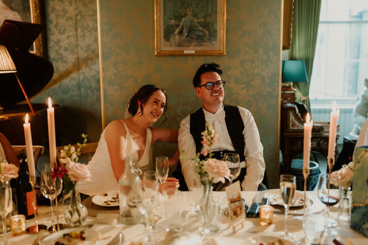 Smiling bride in a white dress and groom in a vest and glasses sit at a candlelit wedding reception table with flowers and glassware.