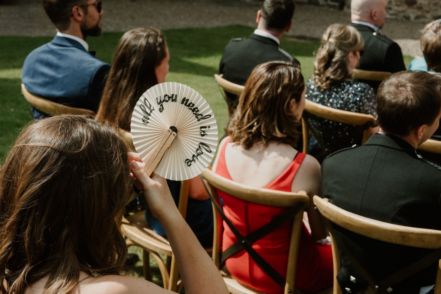 Wedding guests seated on wooden chairs outdoors, with one woman holding a white handheld paper fan with printed text.
