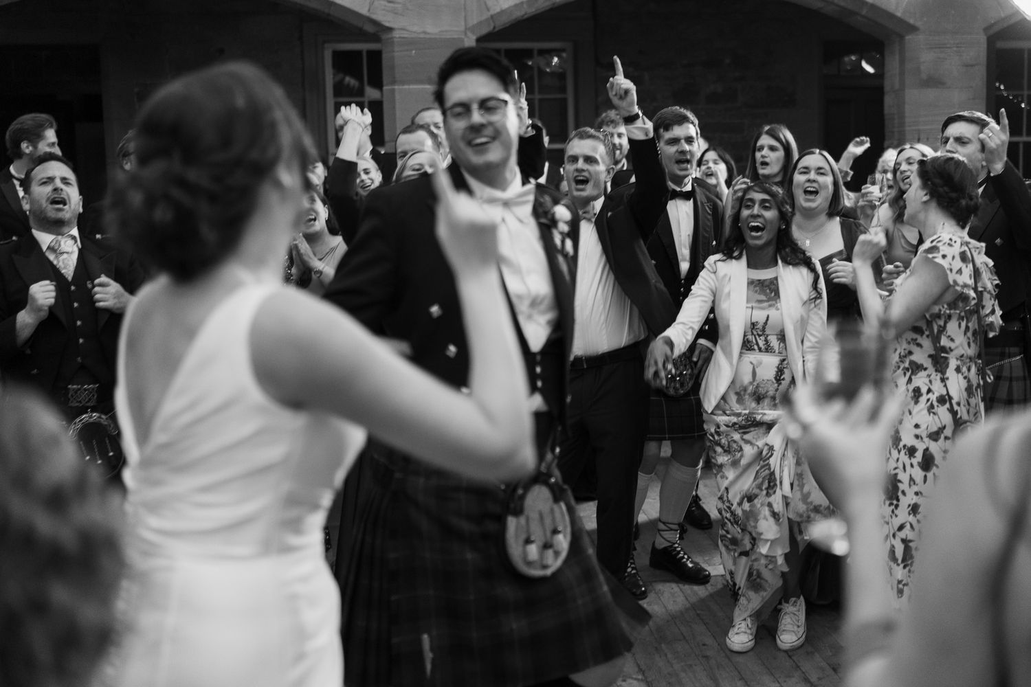 Black and white photo of wedding guests cheering and dancing, with a groom in a kilt at the center of the lively reception.