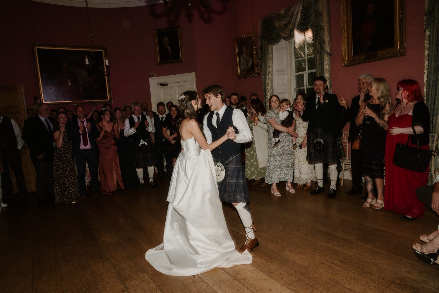 Bride and groom share first dance in elegant pink ballroom surrounded by wedding guests in formal attire.