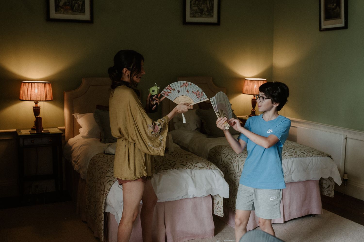 A woman in a gold robe and a boy in blue exchange decorative fans in a warmly lit bedroom.