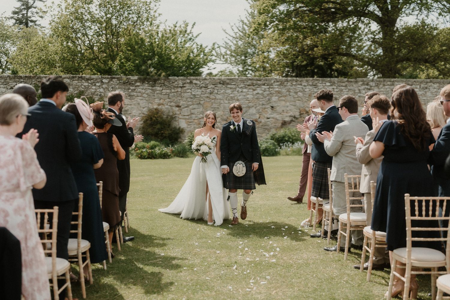 Bride and groom walk down outdoor aisle at Scottish wedding, guests applauding, stone wall backdrop.