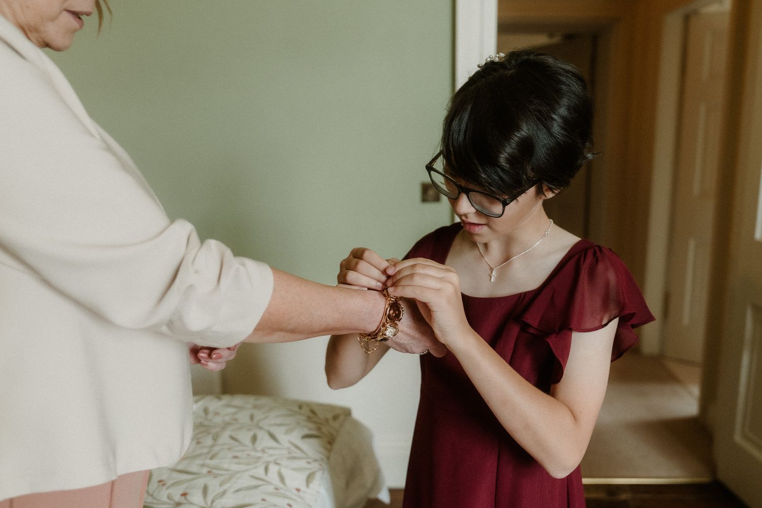 Bridesmaid in burgundy dress fastening bracelet on bride's wrist during wedding preparation.