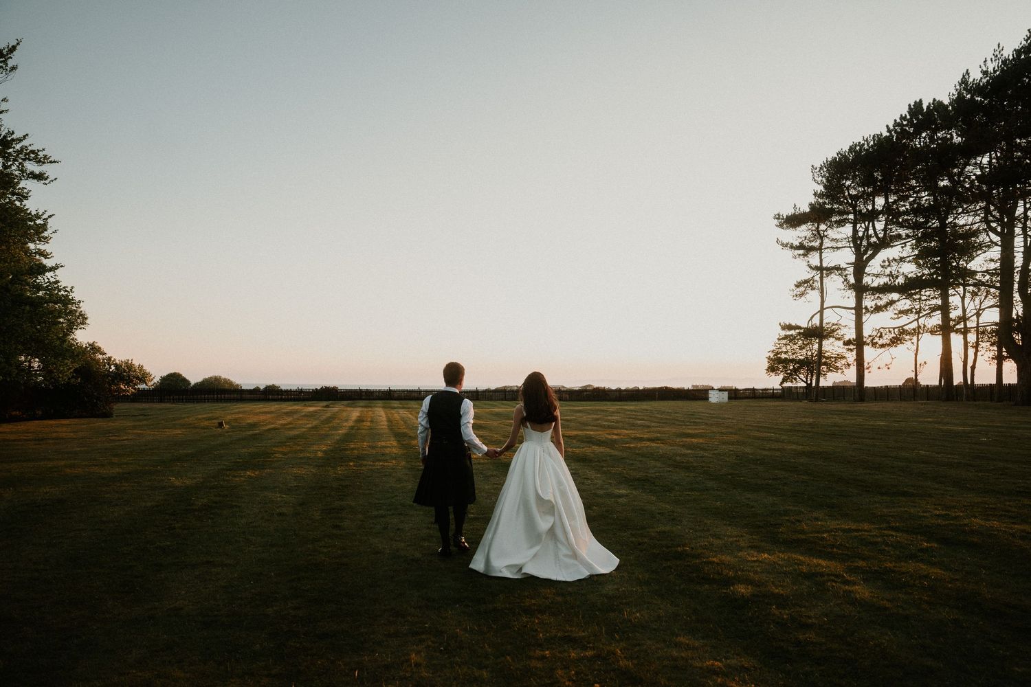 Couple in wedding attire stands in silhouette against a dramatic sunset on a grassy hilltop with pine trees in the distance.