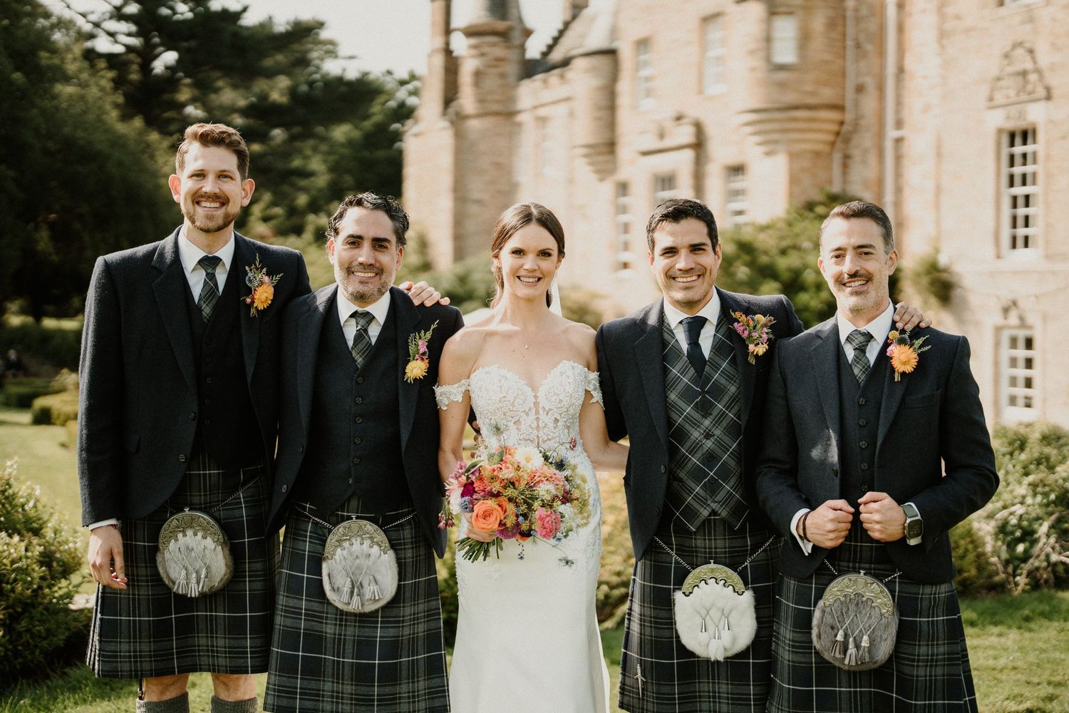 Wedding group stands in front of stone castle wearing traditional Scottish Highland dress.