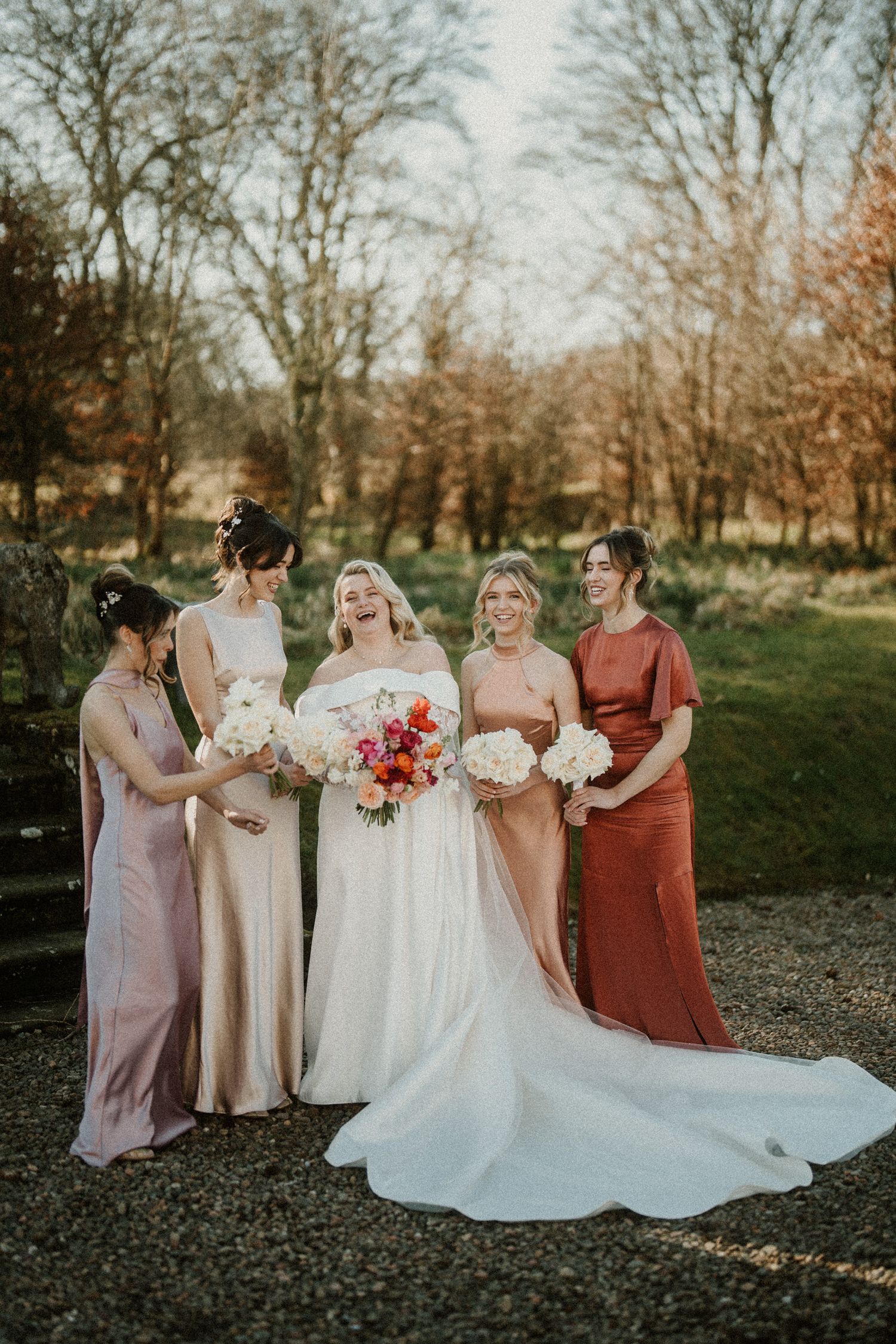 Wedding party shares candid moment in mix of coral and neutral colored dresses outdoors.