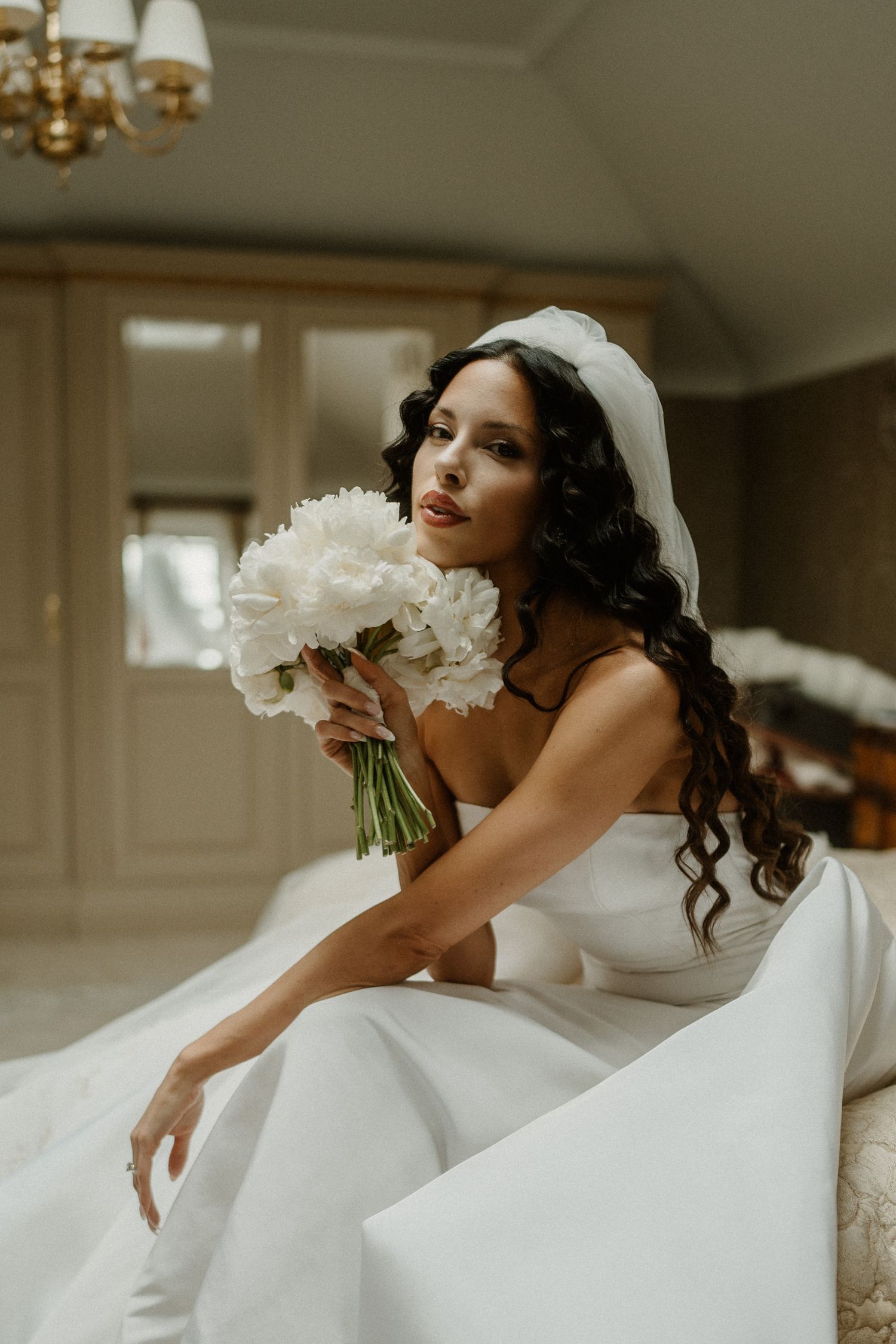 A bride in a white wedding dress holds a bouquet of white flowers while posing elegantly in an indoor setting.