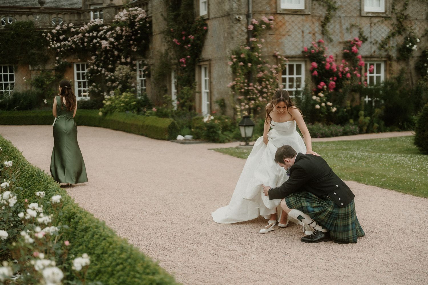 Groom in tartan kilt adjusting bride's shoes on a garden path outside a historic manor house.