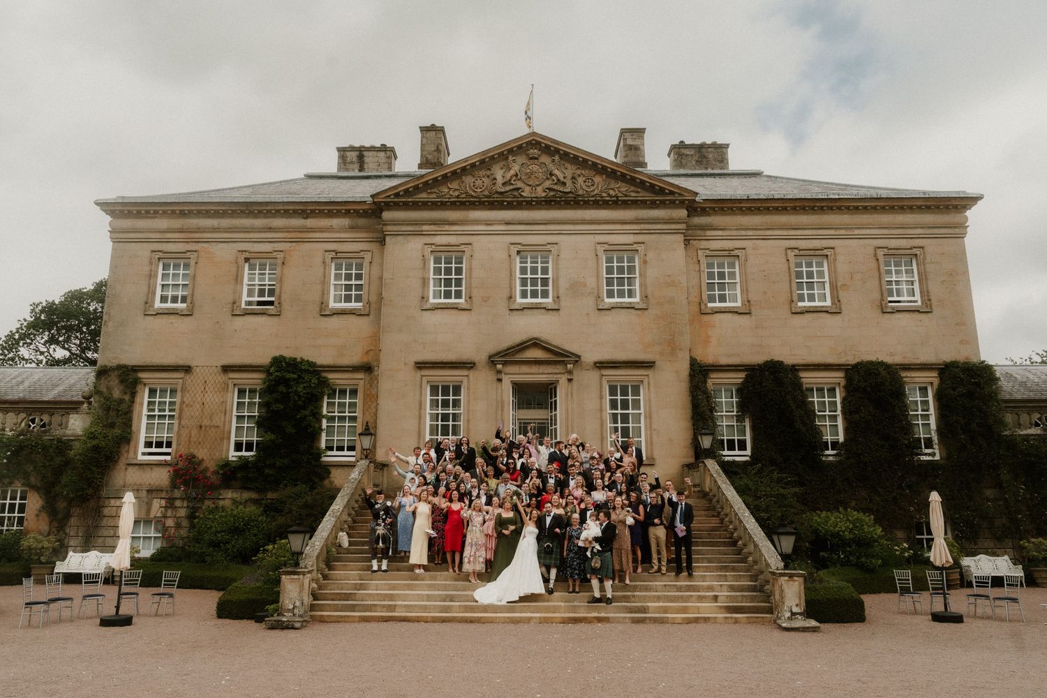 Wedding party posing on grand stone staircase of elegant Georgian manor house venue on cloudy day.