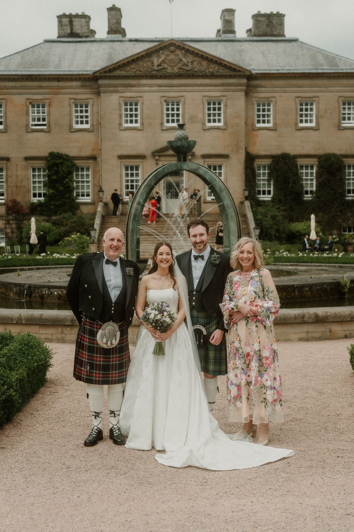 Wedding party posing in front of a Scottish manor house, with groom and father wearing traditional kilts and bride in white gown.