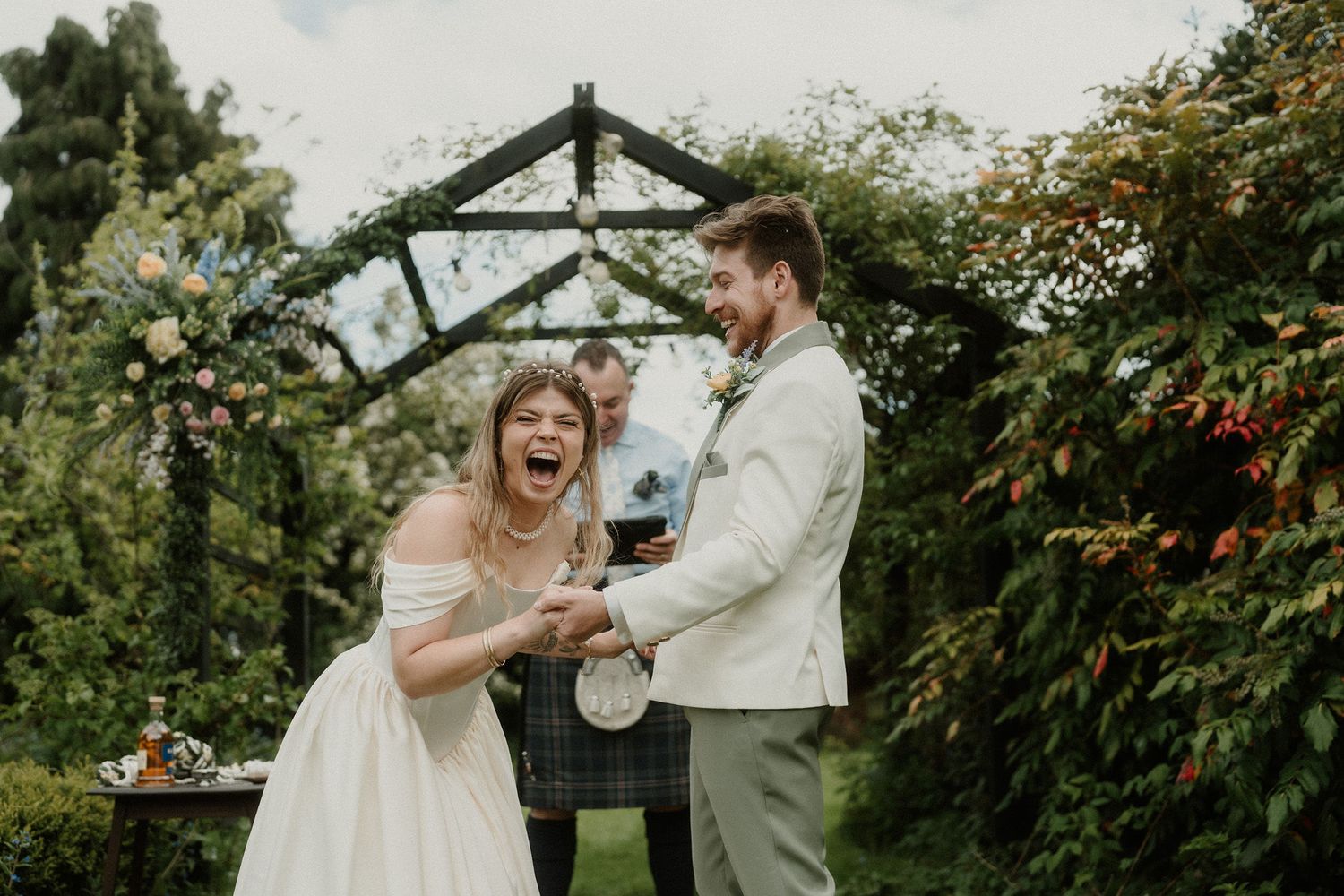 Bride and groom share a joyful laugh during outdoor wedding ceremony beneath a lush green floral arch.