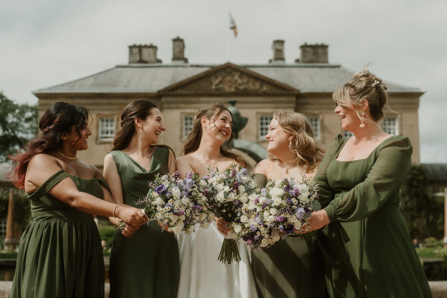 Bride in white gown with four bridesmaids in olive green dresses holding bouquets outside a grand country house.