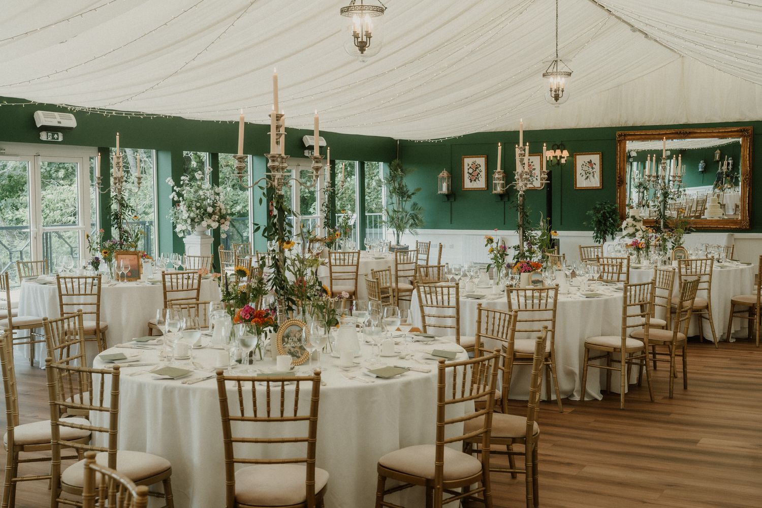 Elegant wedding reception in a marquee with green walls, gold chiavari chairs, white tablecloths, and tall floral centerpieces.