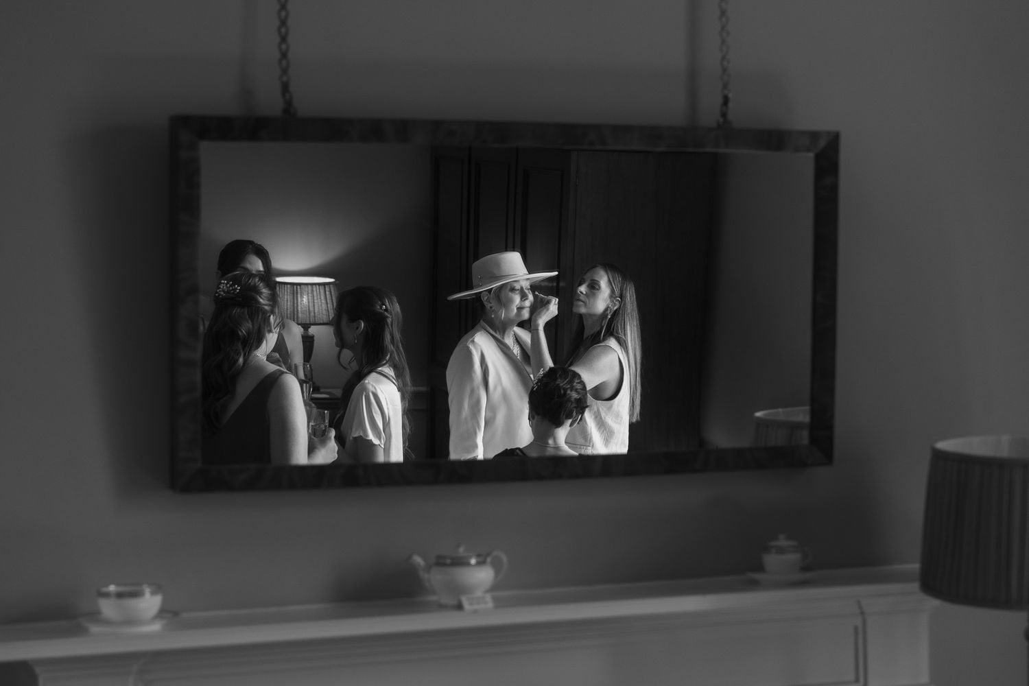 Black and white mirror reflection of a bride getting ready with attendants, one wearing a wide-brim hat.