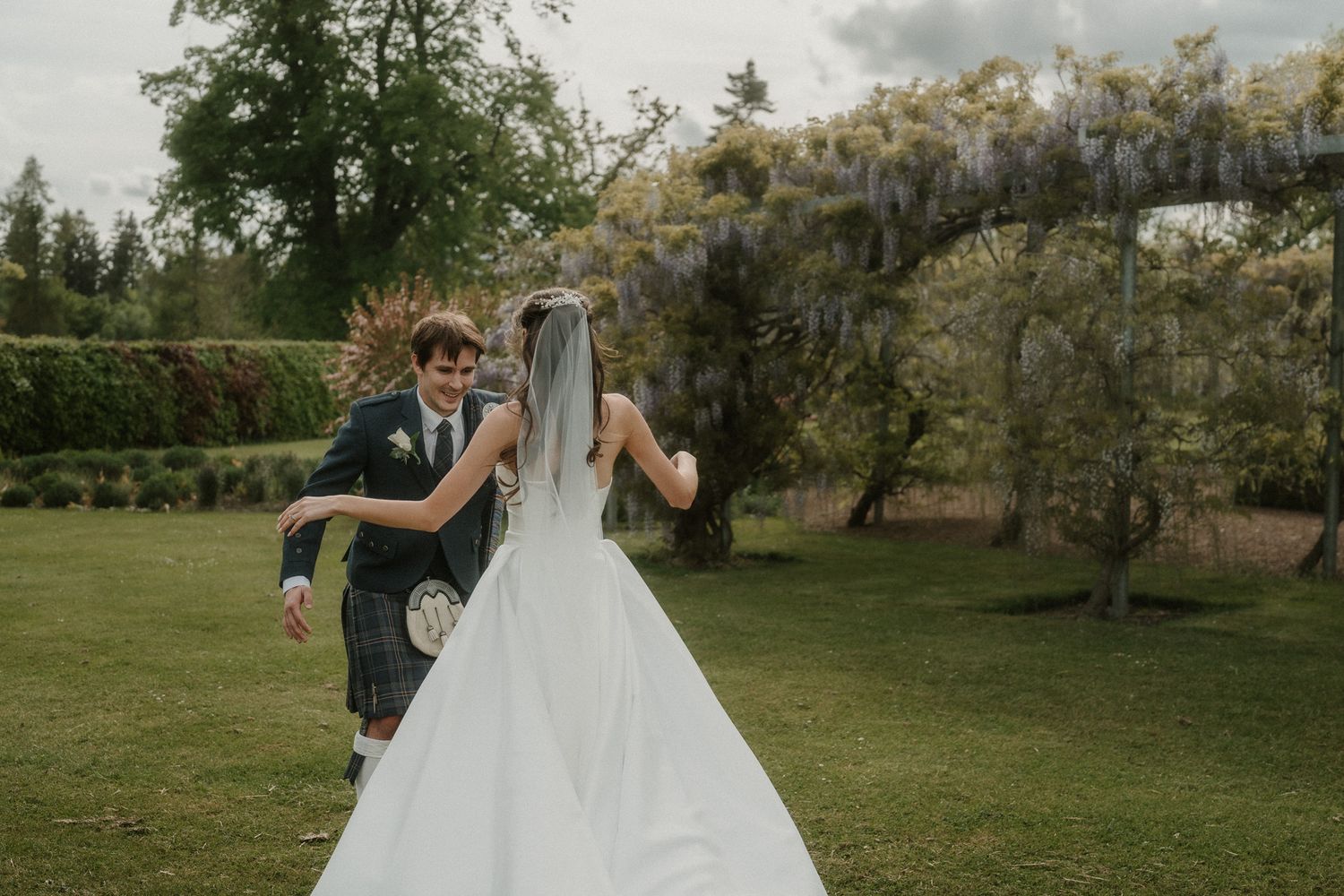 Bride and groom dancing in lush garden with wisteria, groom wearing traditional Scottish kilt on wedding day.
