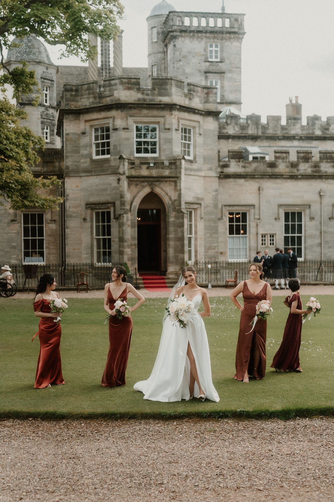 Bride in white gown with bridesmaids in rust dresses posing on lawn before a grand Scottish castle.