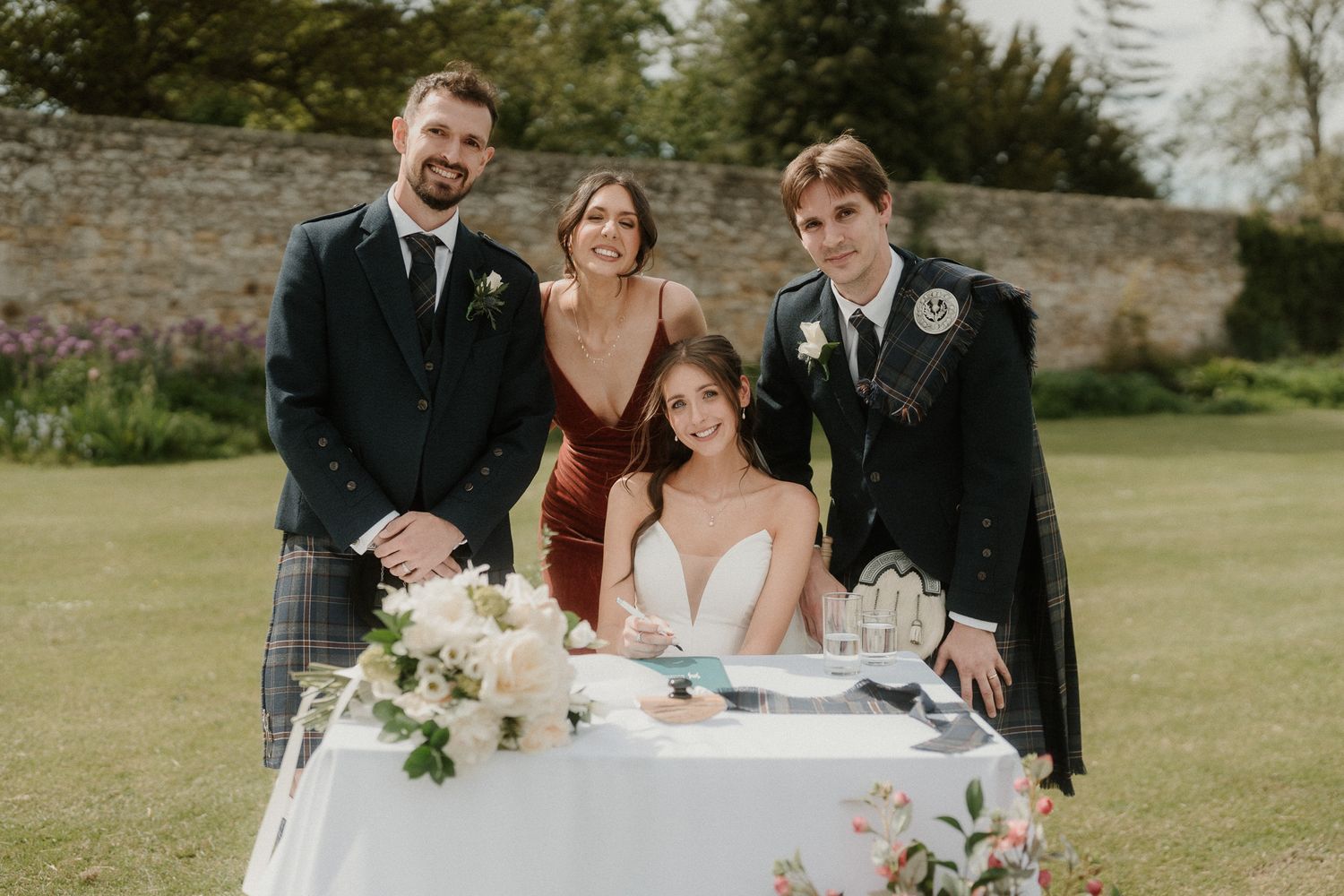 Bride signing wedding register outdoors with groom and guests in Scottish kilts, surrounded by floral arrangements.