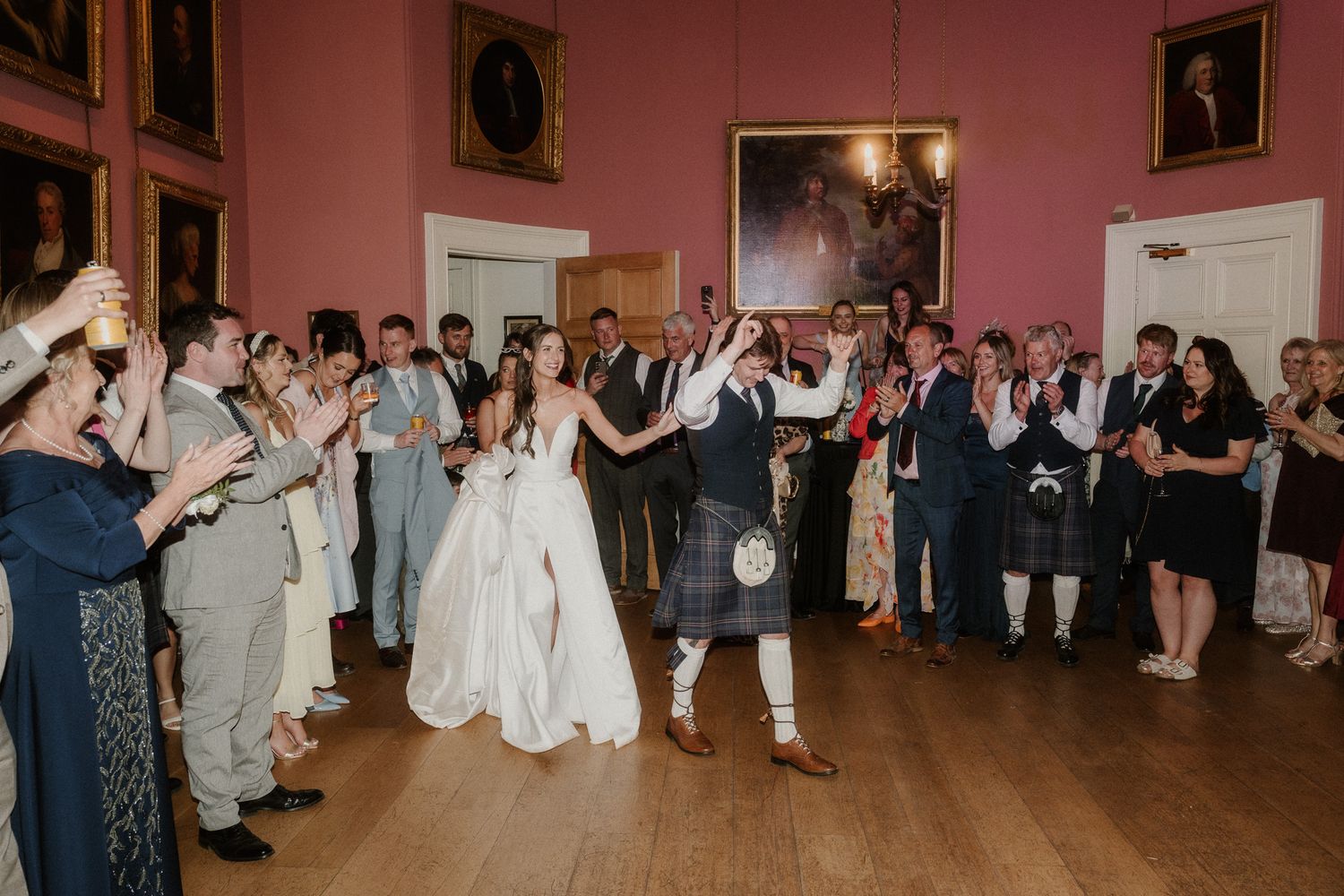 Bride and groom enter wedding reception in elegant pink ballroom, guests applaud as groom wears traditional Scottish kilt.