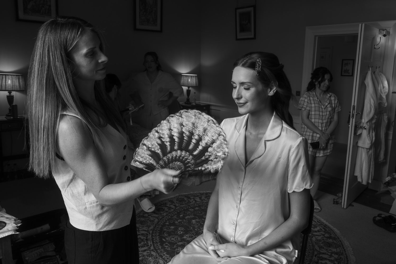 Black and white photo of a woman fanning a seated bride in pajamas while others prepare in the background.