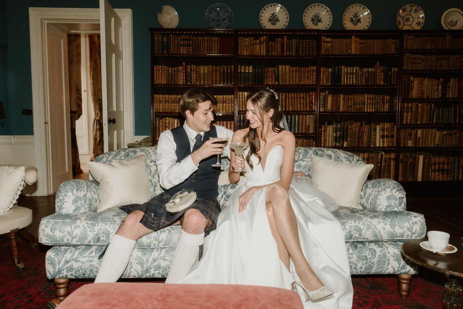 Bride and groom toasting with wine on a floral sofa in an elegant library setting, groom wearing a kilt.
