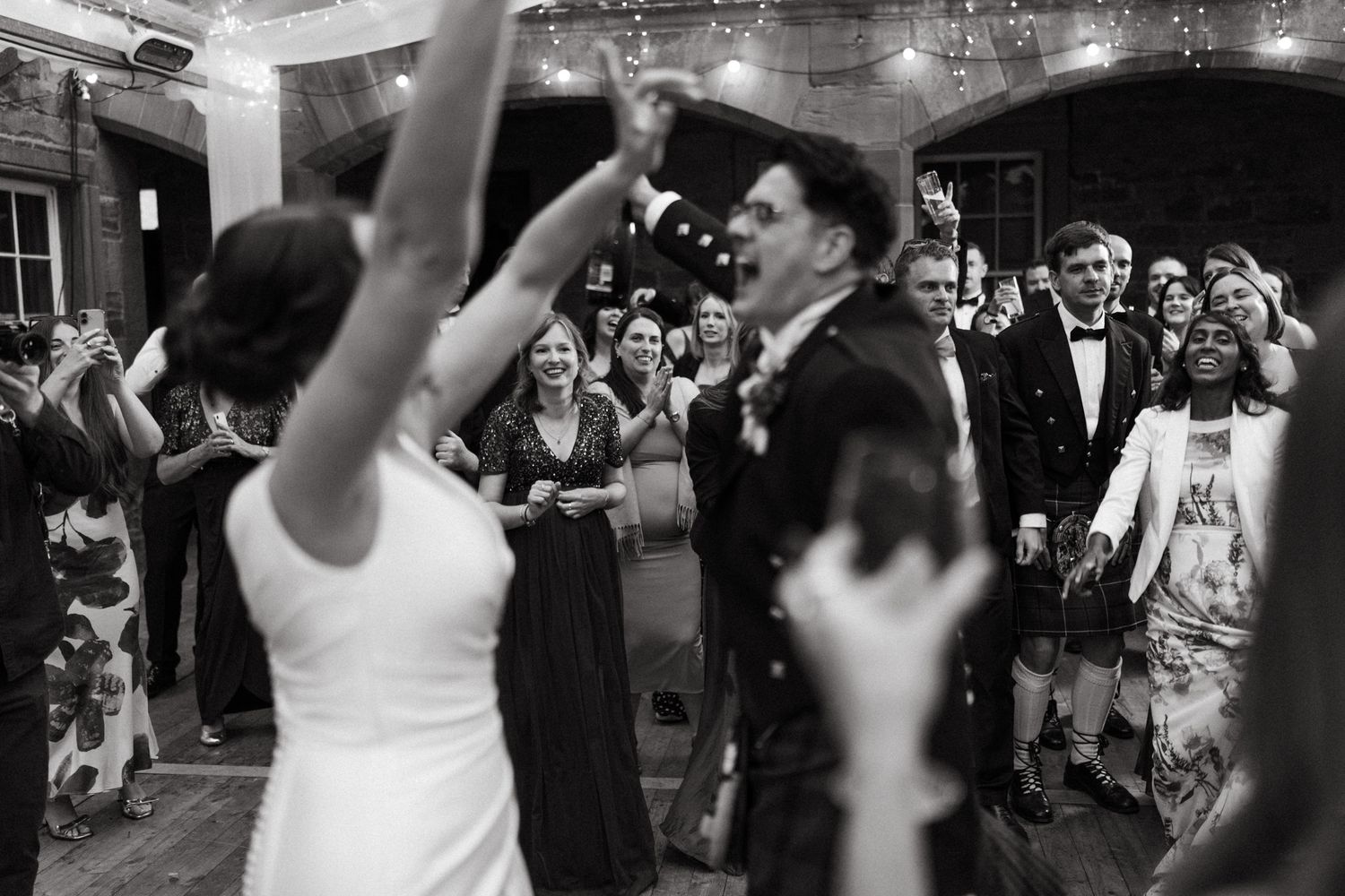 Wedding guests cheer and dance with raised arms as a groom in a dark suit celebrates in a crowded, softly lit reception hall.