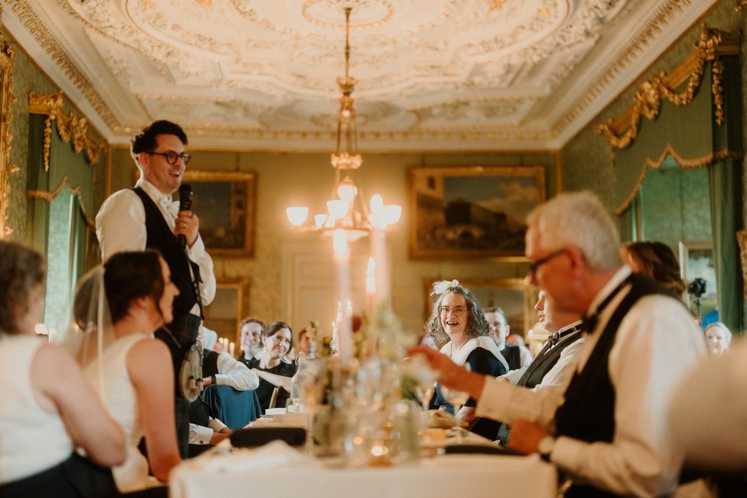 Groom in vest gives a wedding speech holding a microphone as seated guests laugh in an ornate reception hall with a chandelier.