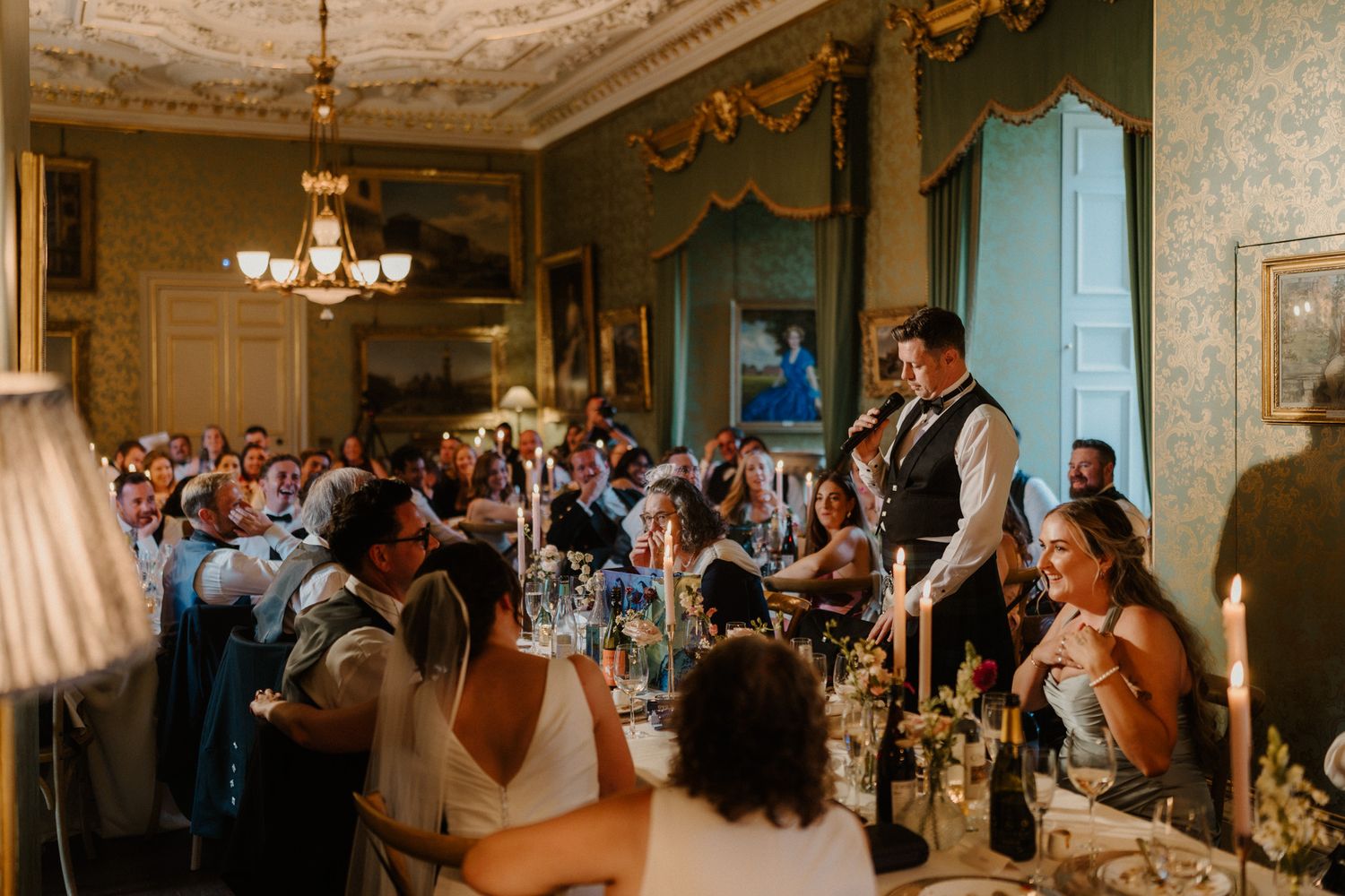 Wedding guests seated at candlelit tables listen to a man in a waistcoat giving a speech in an elegant, chandelier-lit historic ballroom.