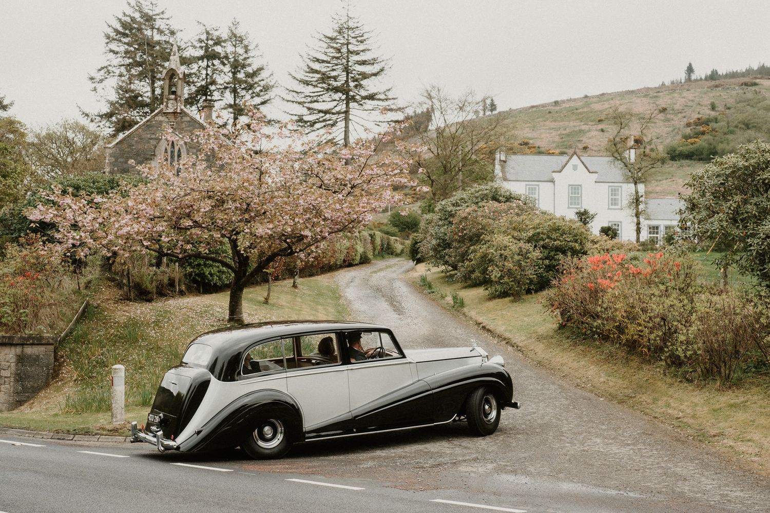 A vintage silver luxury car drives along a scenic coastal road lined with autumn trees and shrubs.