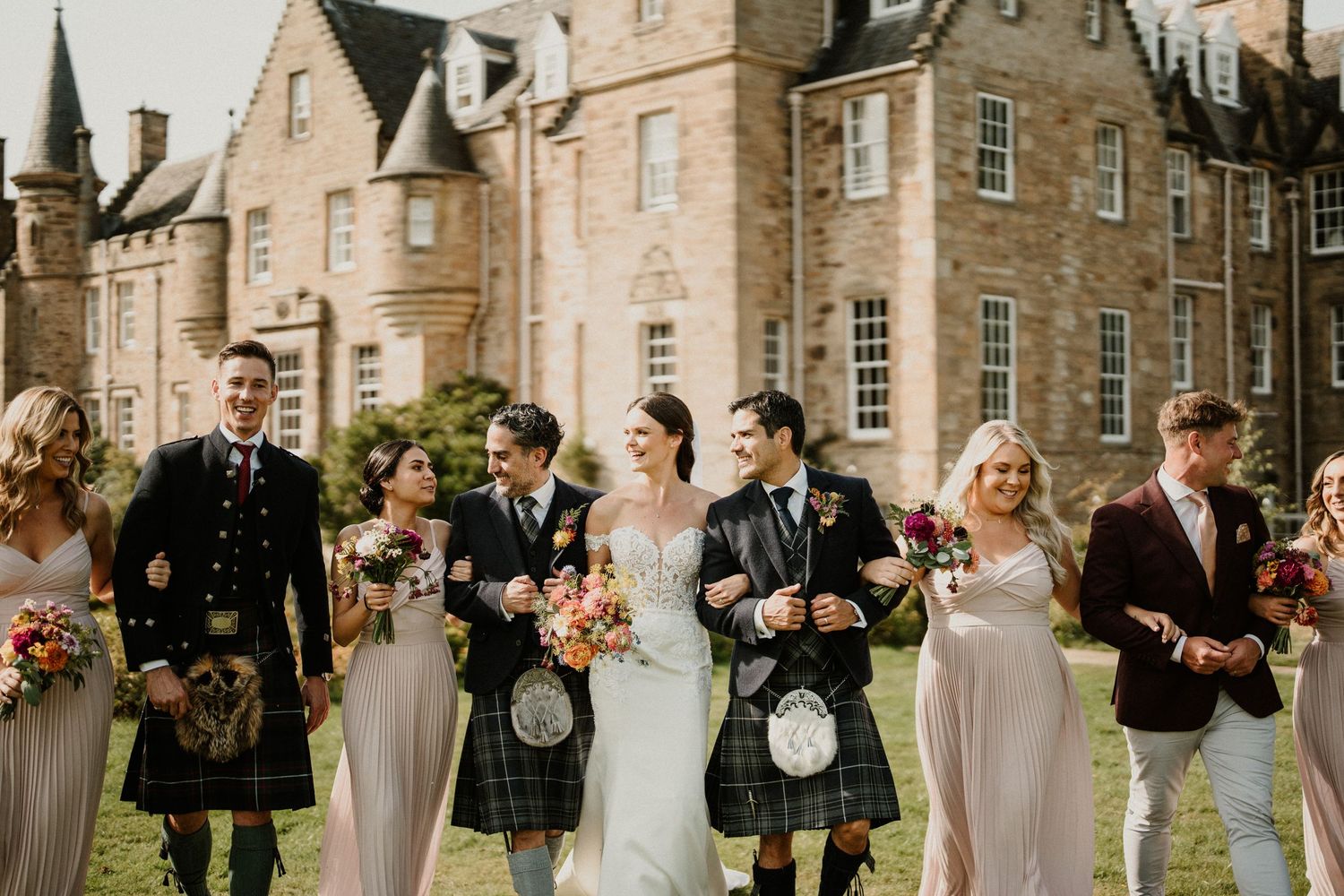 Wedding party poses in front of a grand Scottish castle wearing traditional kilts and formal attire.
