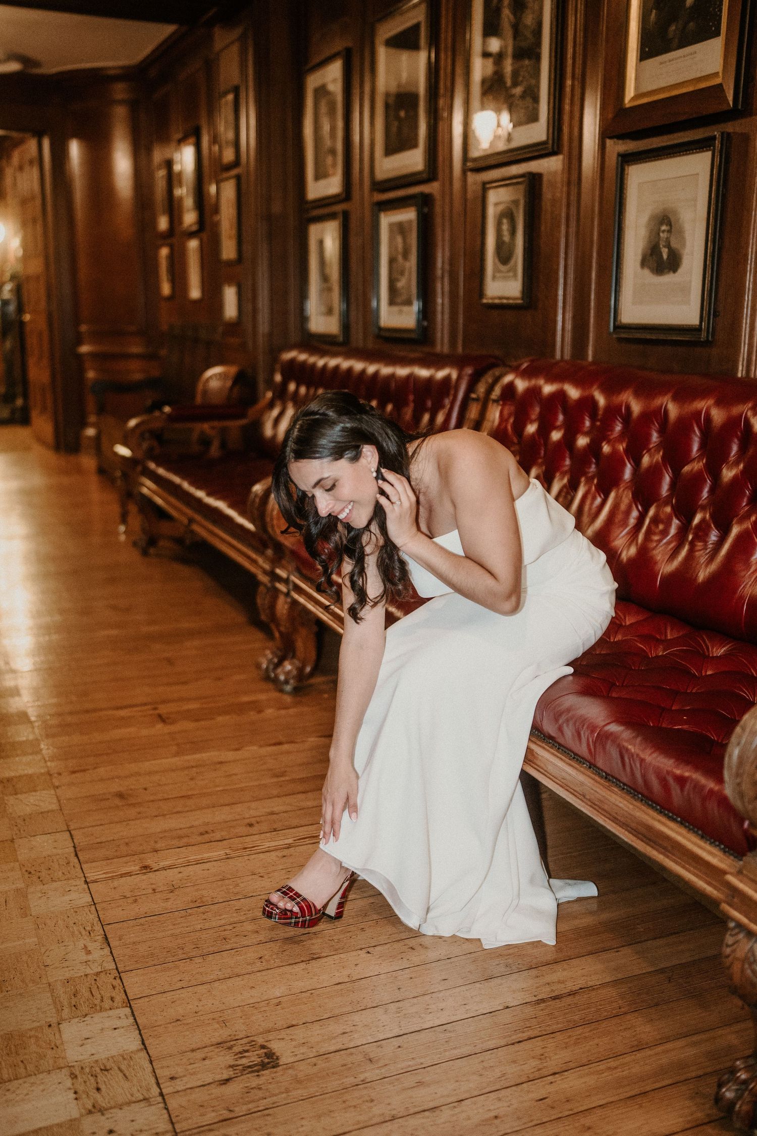 A bride in a white dress sits on a red leather bench in an elegant wood-paneled room with framed portraits.