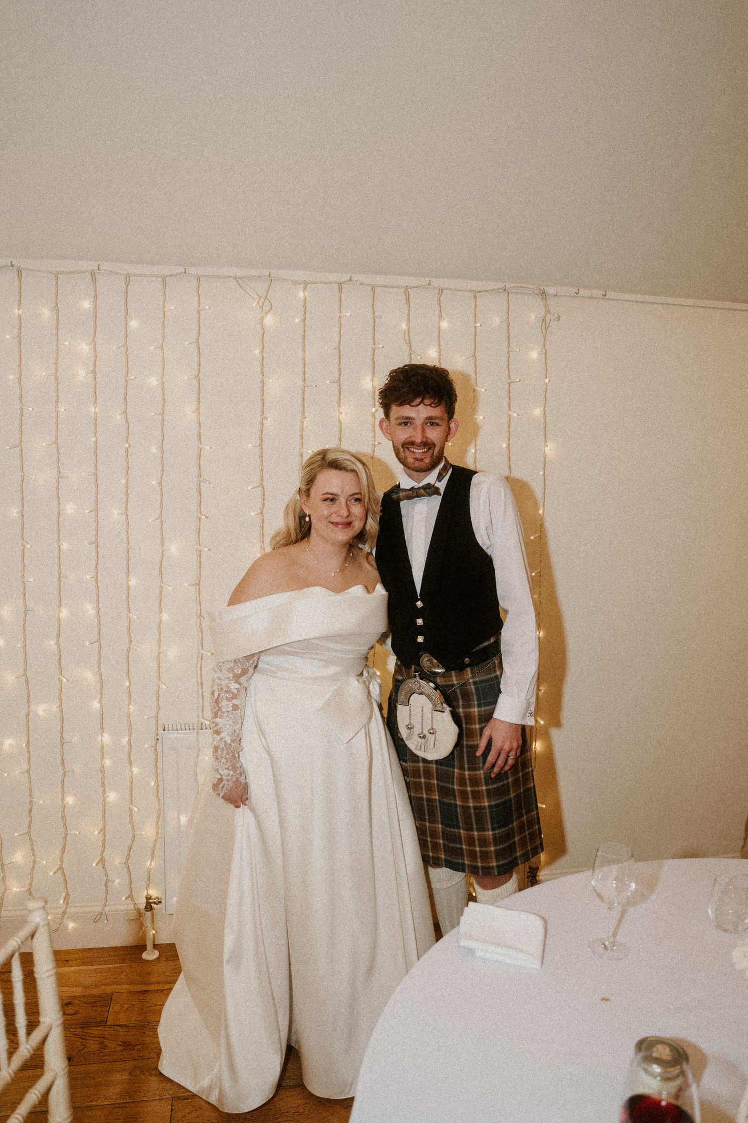 Wedding couple posing against a wall with fairy lights in a casual celebratory moment.