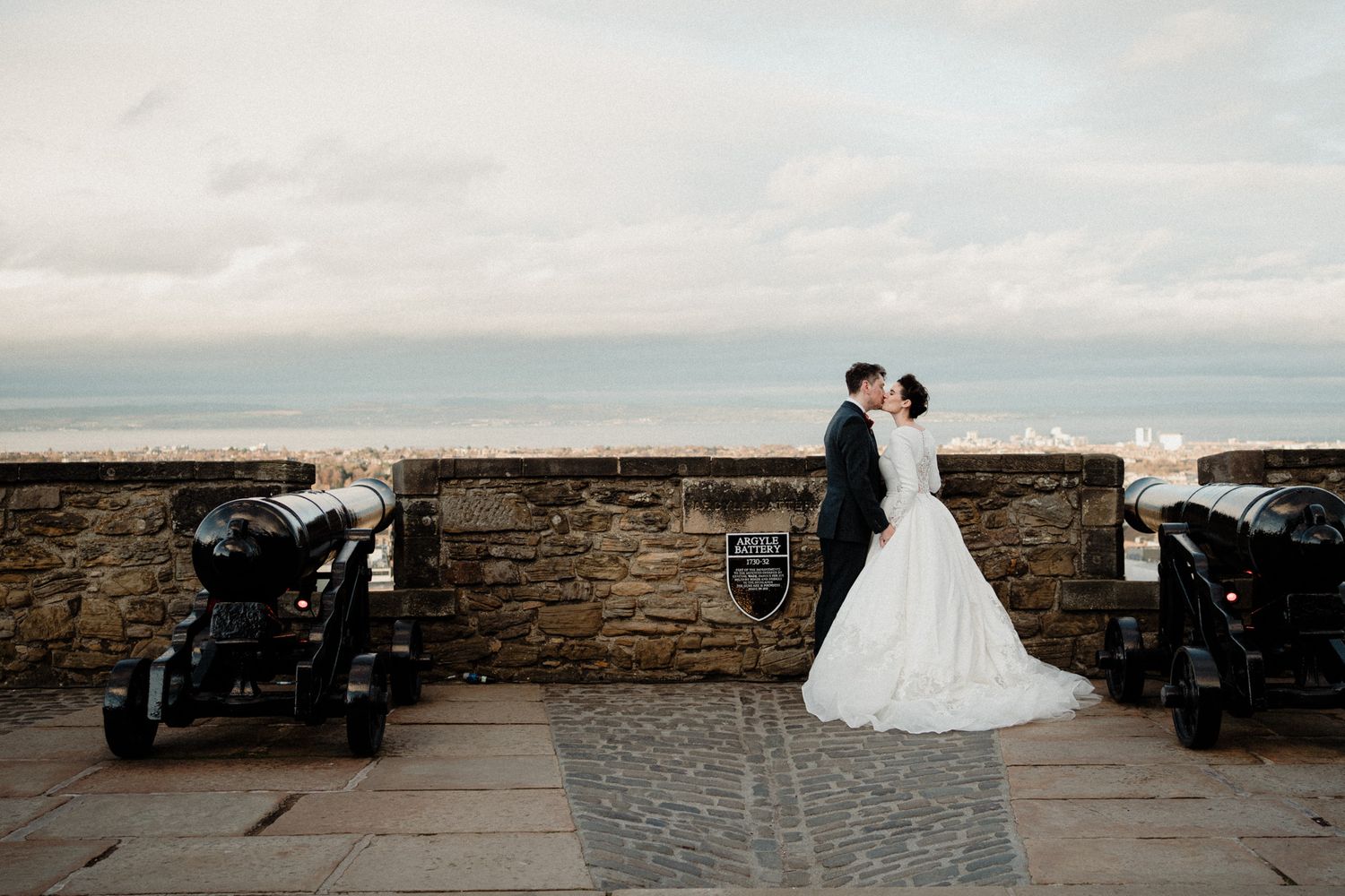 Elegant wedding couple poses between historic cannons on castle battlements.