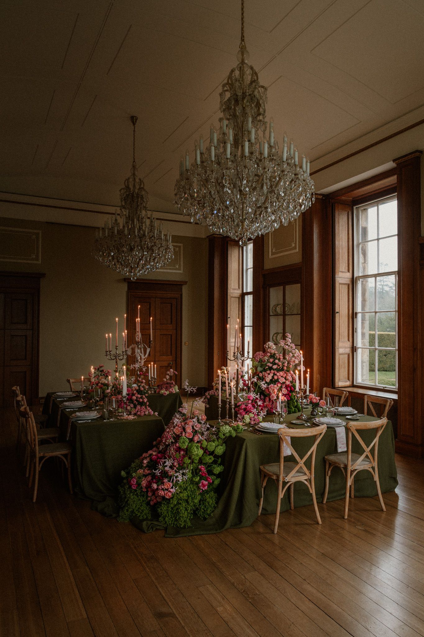 Elegant dining room with crystal chandeliers, wooden floors, and pink floral arrangements on tables with white chairs.