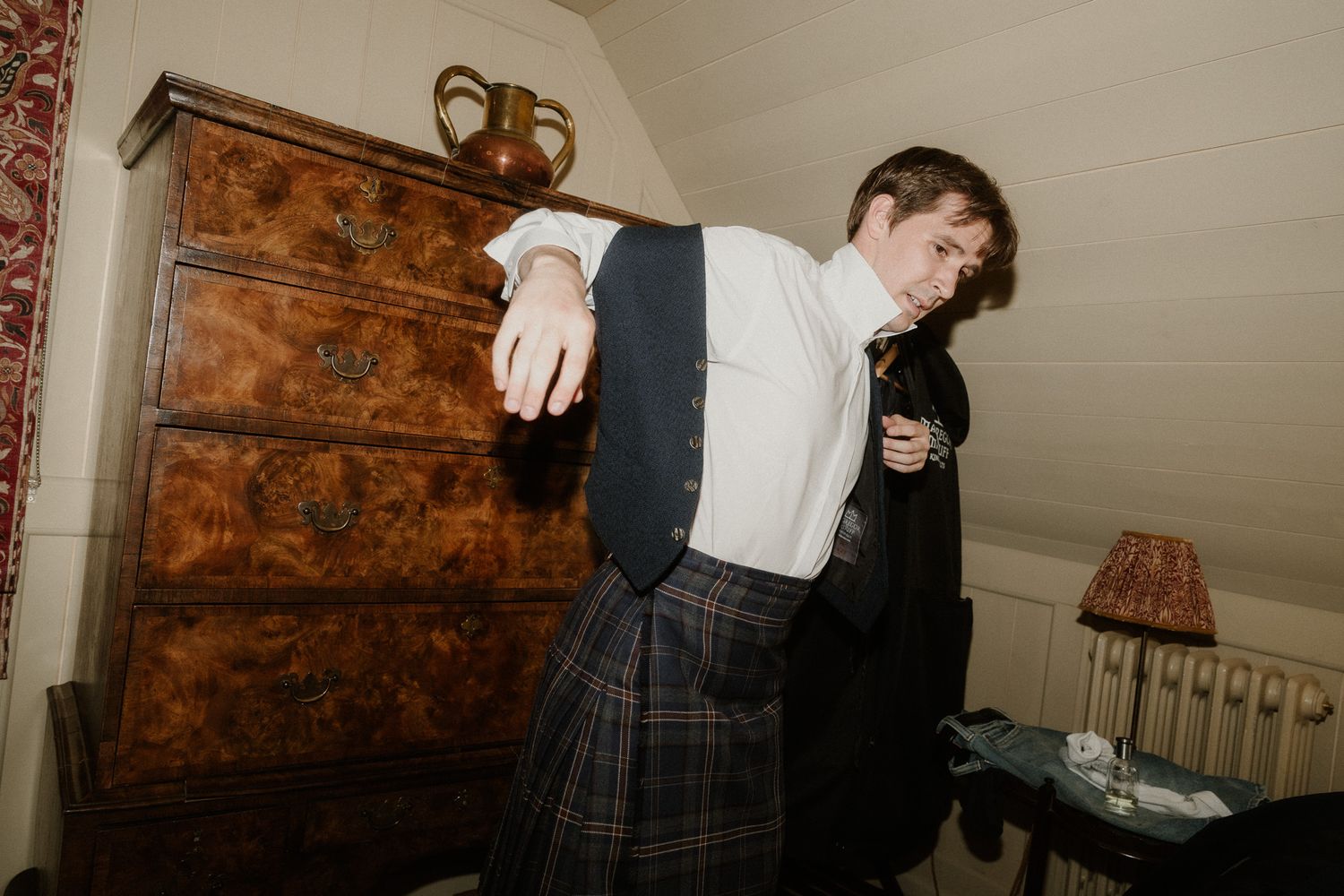 Man in traditional Scottish kilt and waistcoat dancing indoors near antique wooden dresser.