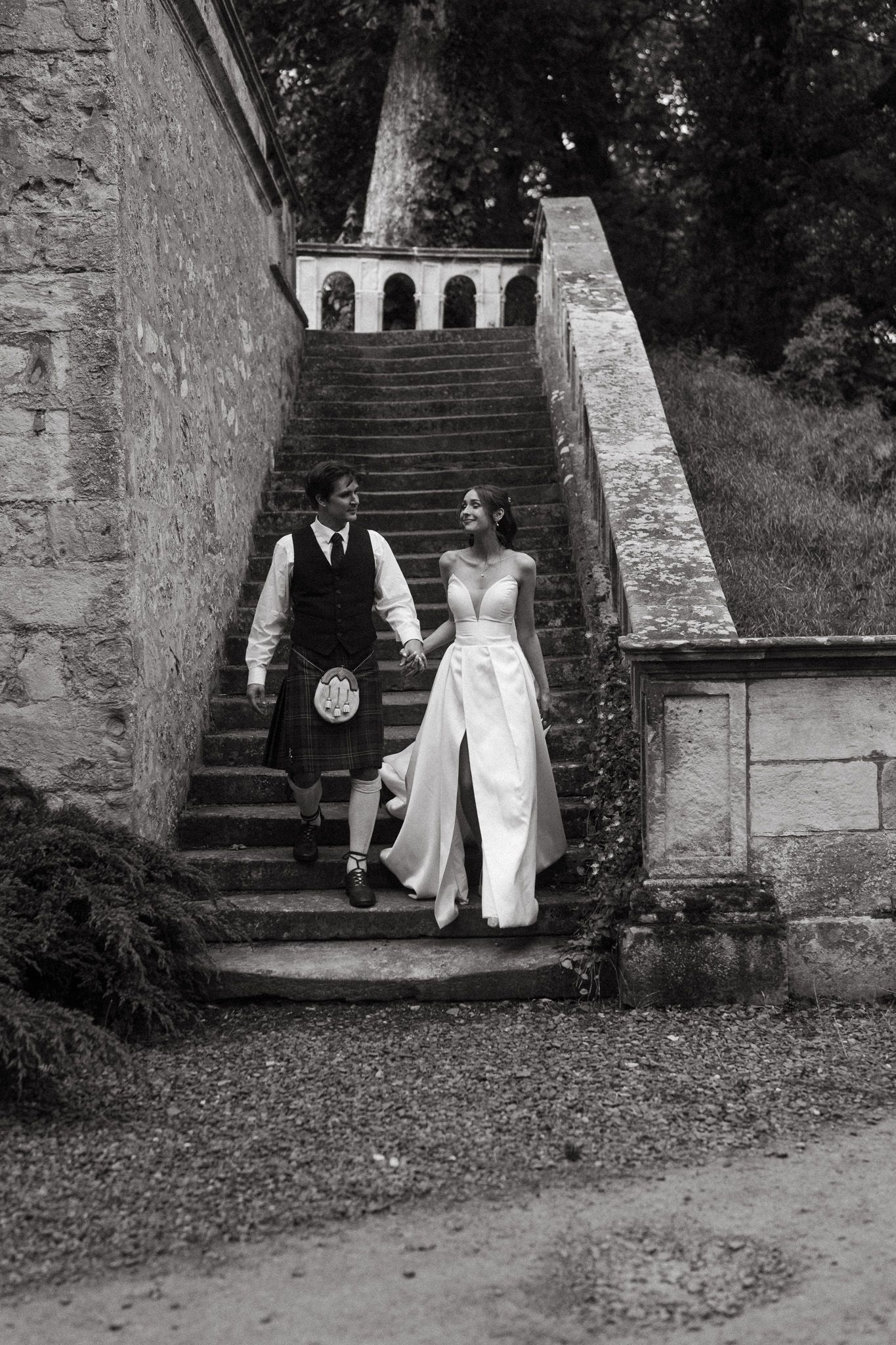 Black and white photo of a bride and groom holding hands walking down stone stairs at a castle wedding venue.