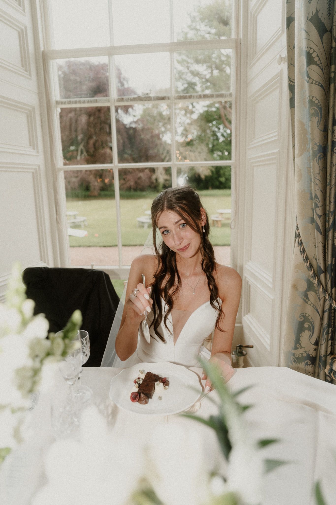 A bride in a white gown smiles while enjoying dessert at her wedding reception table by a large window.