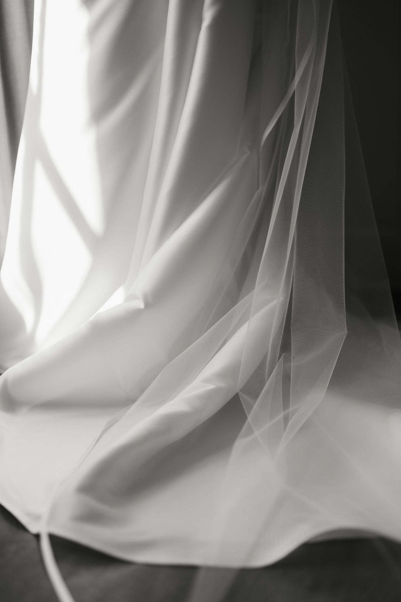 Black and white photo of a sheer tulle wedding veil draped over soft satin fabric beside a sunlit window.