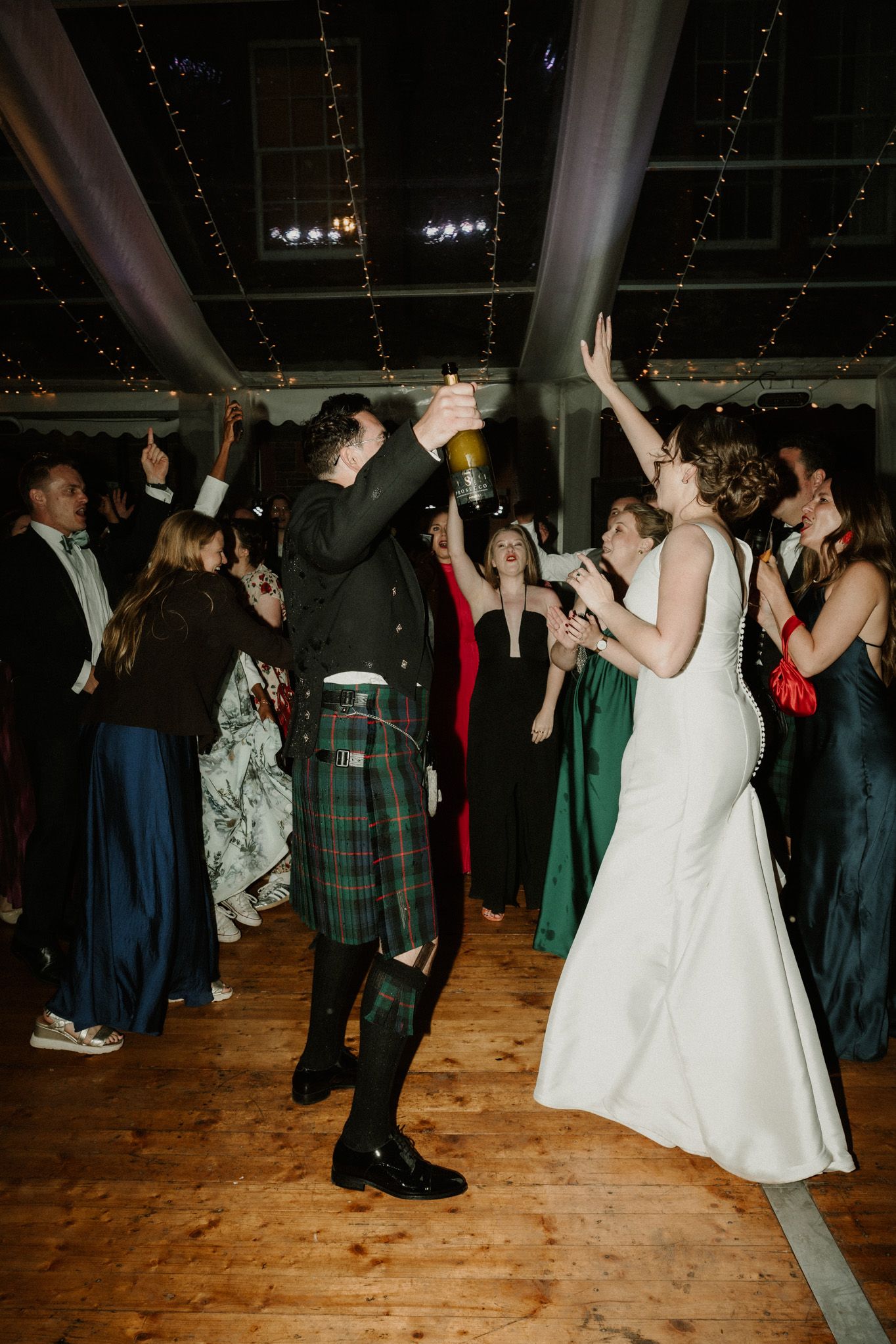 Groom in a green tartan kilt raises a champagne bottle while dancing with the bride in a white gown and guests at a wedding reception.