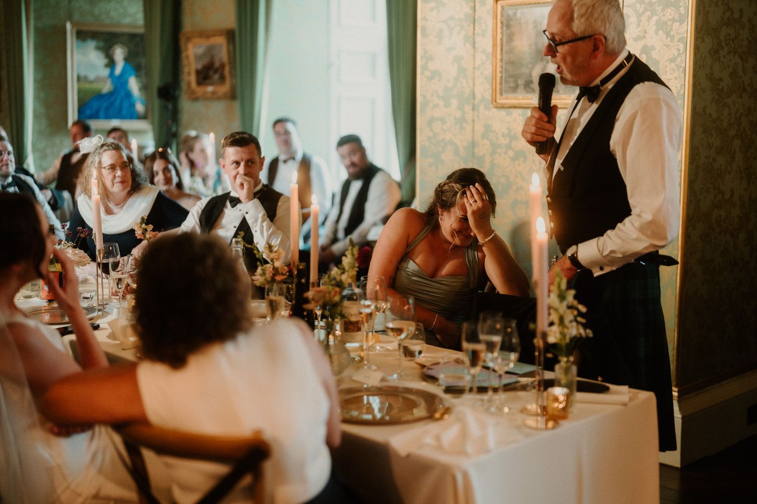 Man in a vest and bow tie gives a wedding toast with a microphone while seated guests laugh at candlelit banquet tables.
