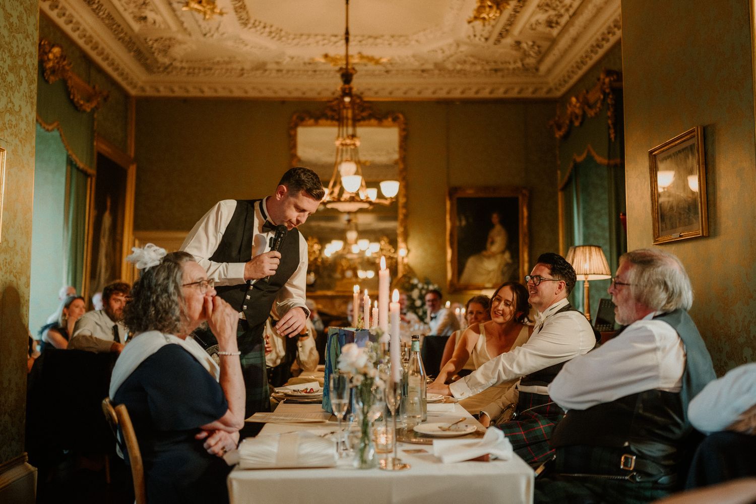 Waiter in a vest pours wine for guests seated at a candlelit dinner table in an elegant dining room with ornate ceilings and chandeliers.