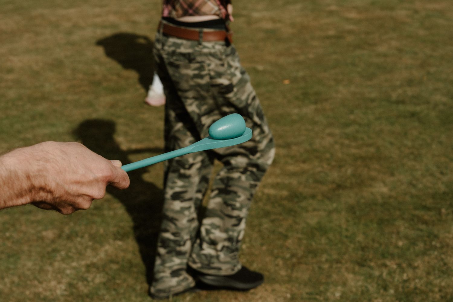 Person in camouflage pants stands on grass while a hand holds out a teal plastic spoon, as in an egg and spoon race.