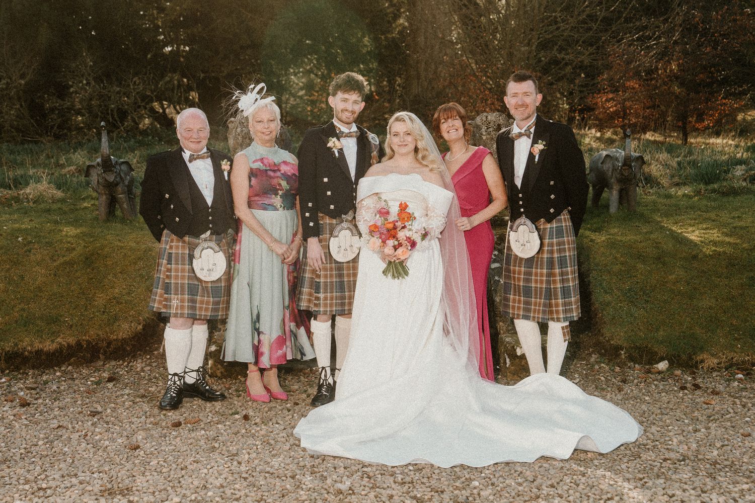 Wedding party poses outdoors in traditional Scottish attire and formal wear.